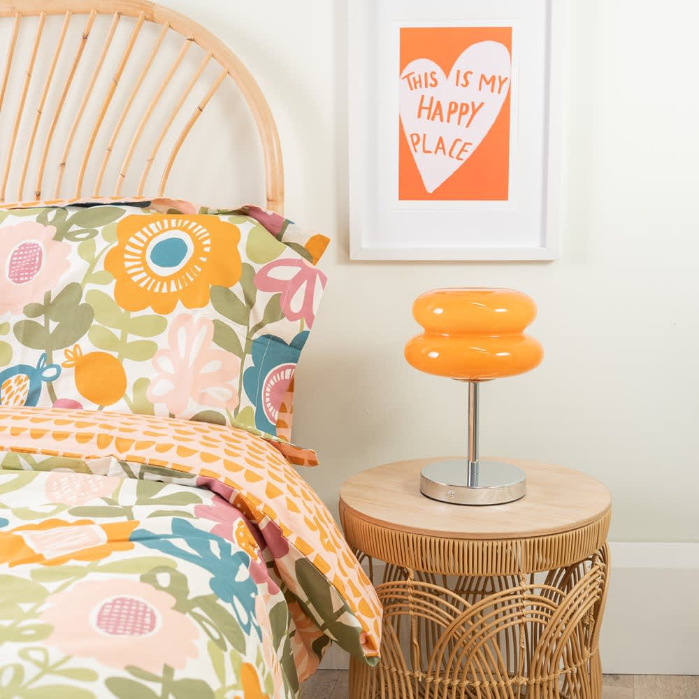 Bedroom with rattan headboard, colorful floral bedding, and an orange retro-style table lamp on a round wicker side table, with framed artwork reading &lsquo;This is my happy place&rsquo; on the wall.