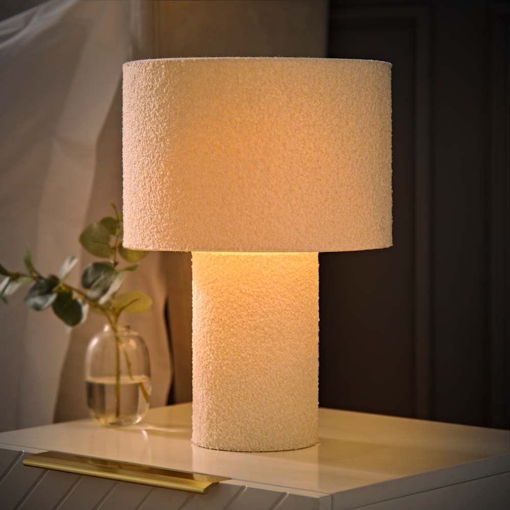 Cream boucle table lamp with a textured cylindrical base and matching shade, softly glowing on a bedside cabinet with a small glass vase beside it.