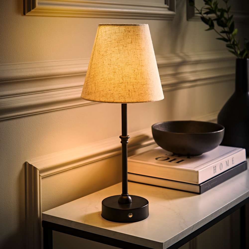 Slim black table lamp with a warm fabric shade glowing on a hallway console table, styled with books, a decorative bowl and a black vase for a cosy modern look.