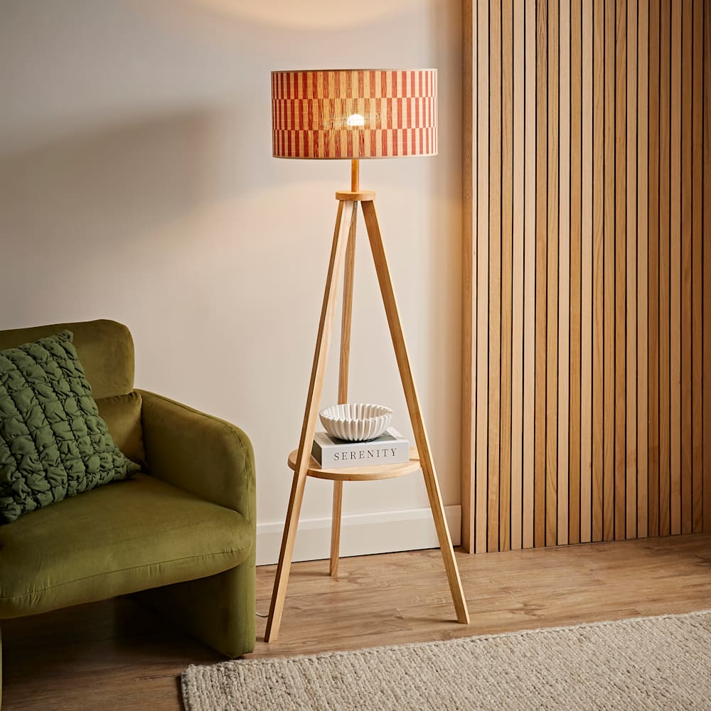 Wooden tripod floor lamp with a built-in circular shelf styled with a book and decorative bowl, topped with a bold red and orange striped drum shade, positioned in a contemporary living room with an olive green velvet sofa and wooden slatted wall panel