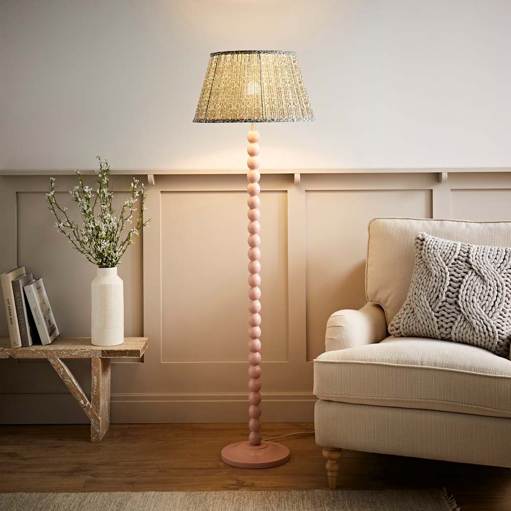 Tall pink bobbin floor lamp with a stacked ball wooden base and a floral pleated shade, styled in a warm neutral living room with a cream armchair, chunky knit cushion, rustic wooden bench and white vase of spring blossom against a taupe panelled wall
