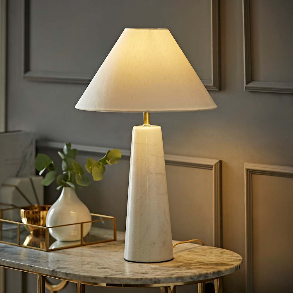 Modern table lamp with a tall, tapered marble-effect base and a cream conical fabric shade, placed on a marble console table with a decorative tray and small vase of greenery against a paneled wall.