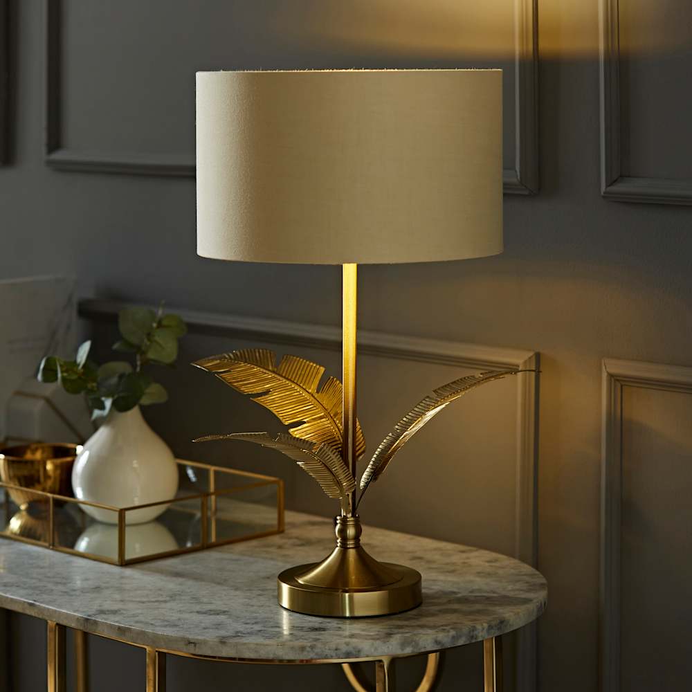 Glamorous gold table lamp with decorative botanical leaf detailing on the stem and a cream drum shade, glowing warmly on a marble and gold console table styled with a white ceramic vase, eucalyptus stems and a gold geometric tray, against a dark grey panelled wall