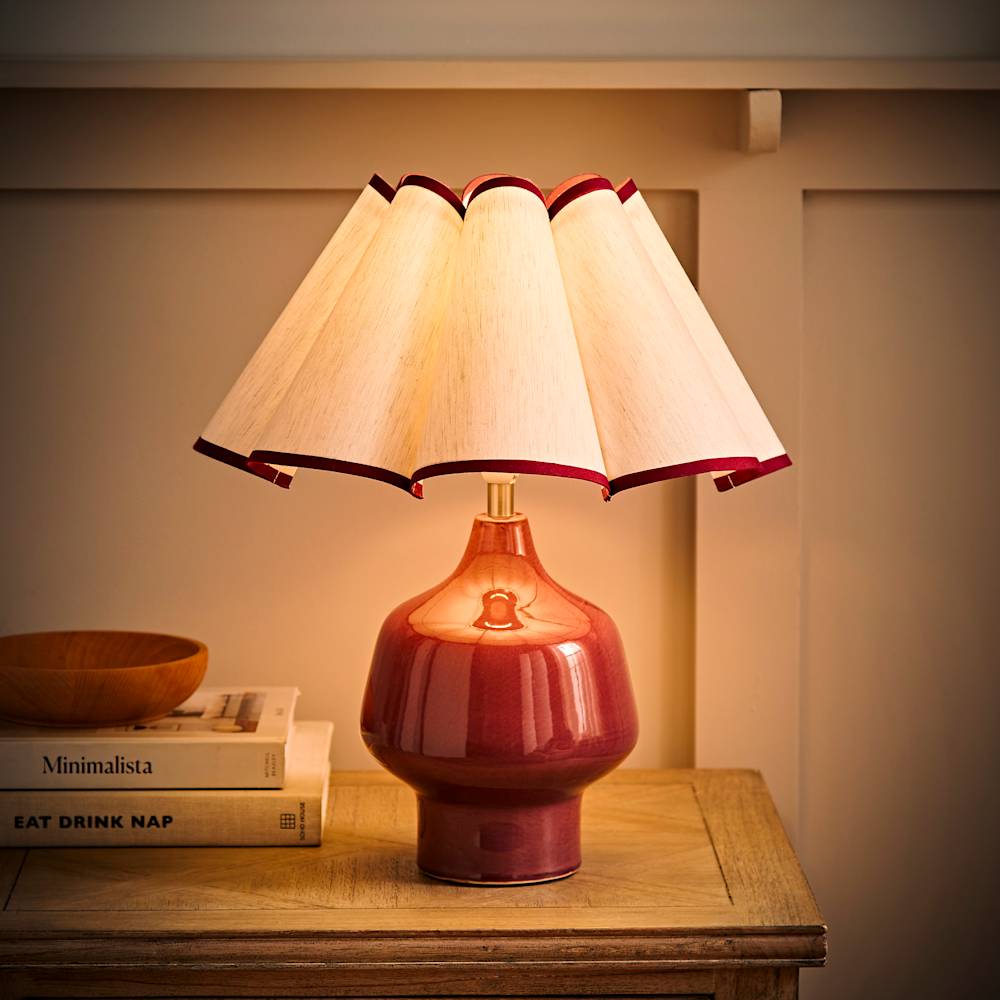Red ceramic table lamp with a scalloped cream lampshade and subtle red trim, styled on a wooden side table with books for a warm, cosy glow.