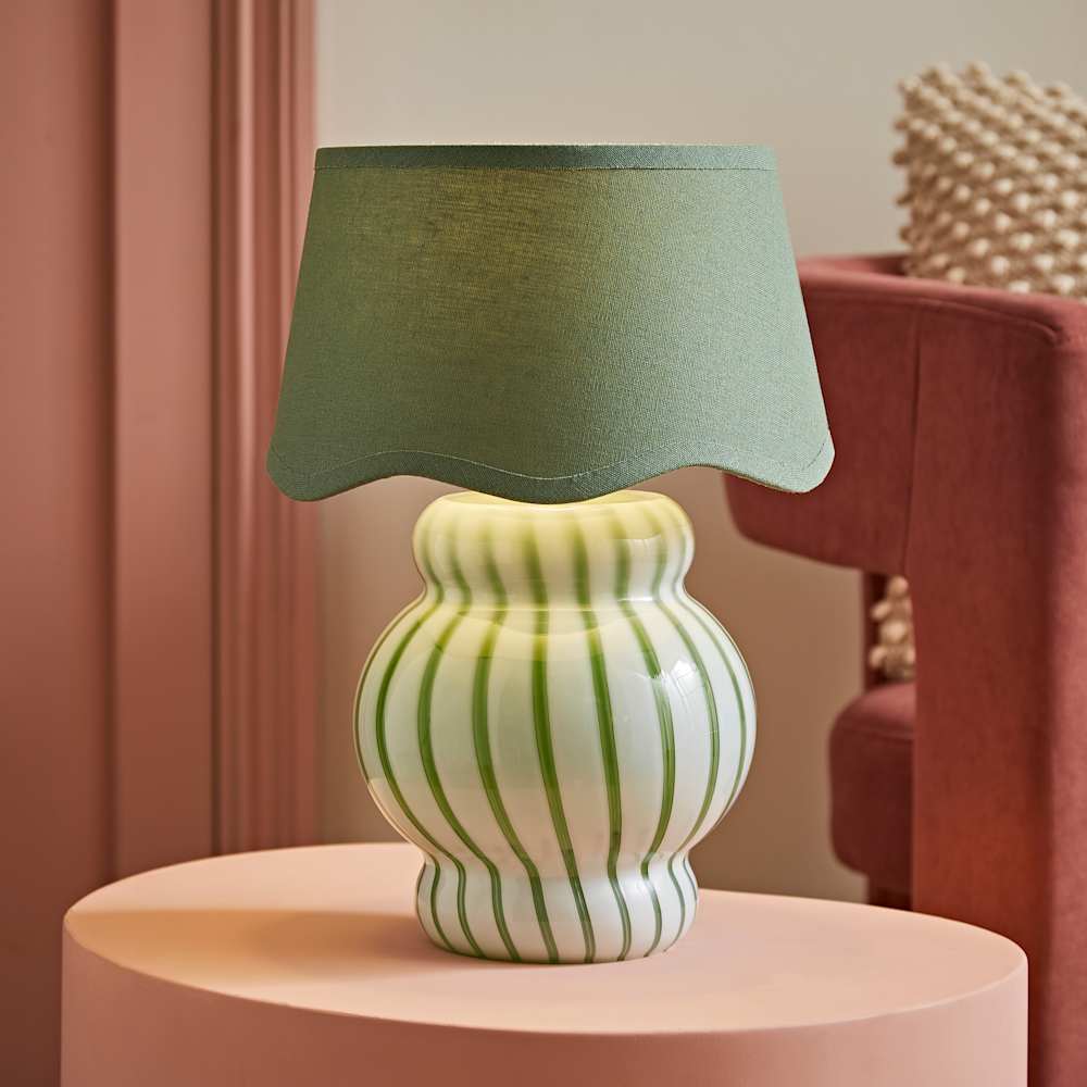 Sculptural statement table lamp with a rounded white ceramic base featuring green stripe detailing and a matching sage green scallop-edged fabric shade, styled on a blush pink plinth against a dusky pink backdrop with a terracotta velvet chair in the background