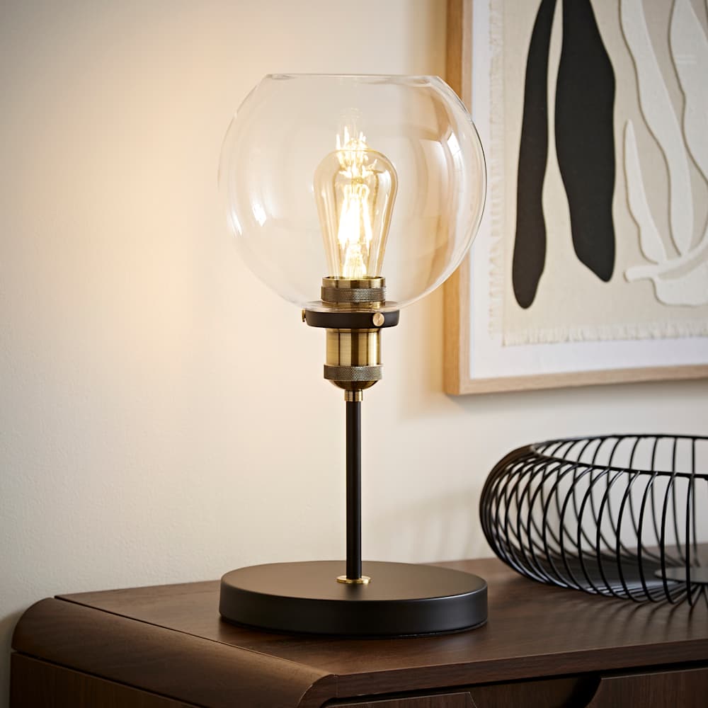Industrial-style table lamp with a round black base, slim black stem, and clear glass globe shade showcasing an exposed filament bulb, placed on a wooden surface with decorative wire bowl and abstract wall art in the background.