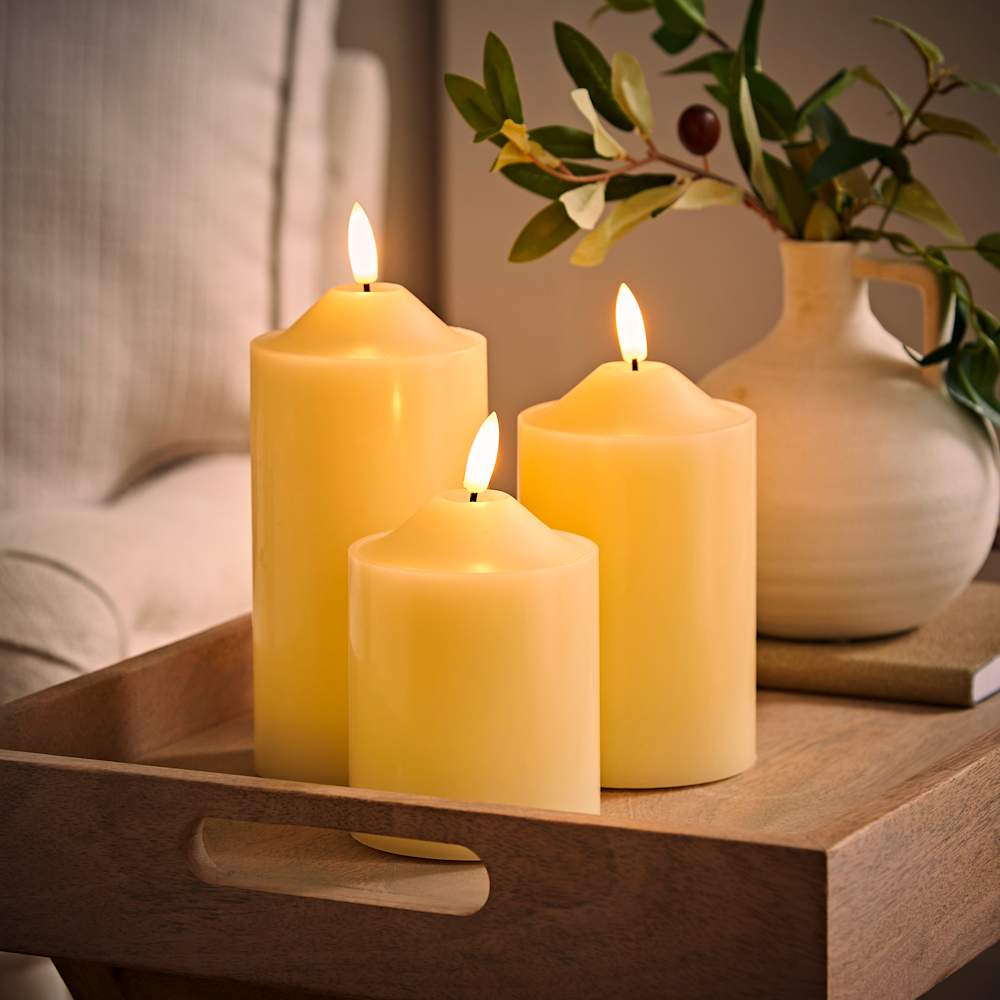 Set of three warm LED pillar candles glowing softly on wooden tray beside ceramic vase - realistic flameless candlelight