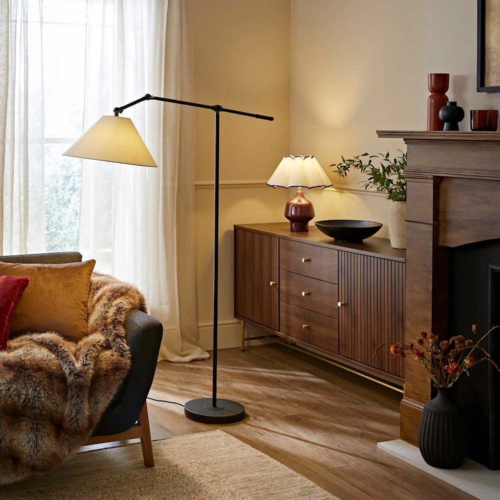 A cosy and warm looking living room with a sofa covered in blankets, dark wood sideboard, a black floor lamp and a burgundy table lamp