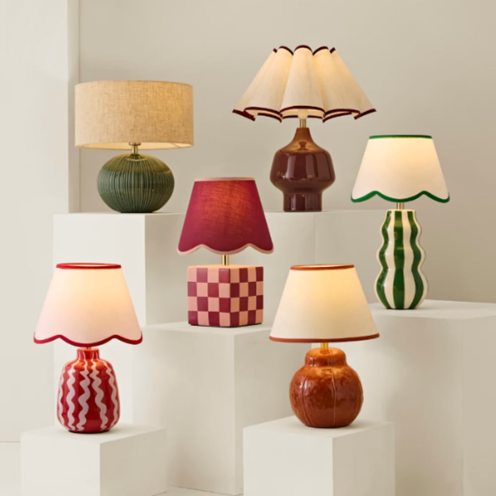 Collection of decorative table lamps displayed on white plinths. Designs feature bold patterns, scalloped shades, and vibrant colors including red, green, and cream, creating a playful, modern aesthetic for home lighting décor.
