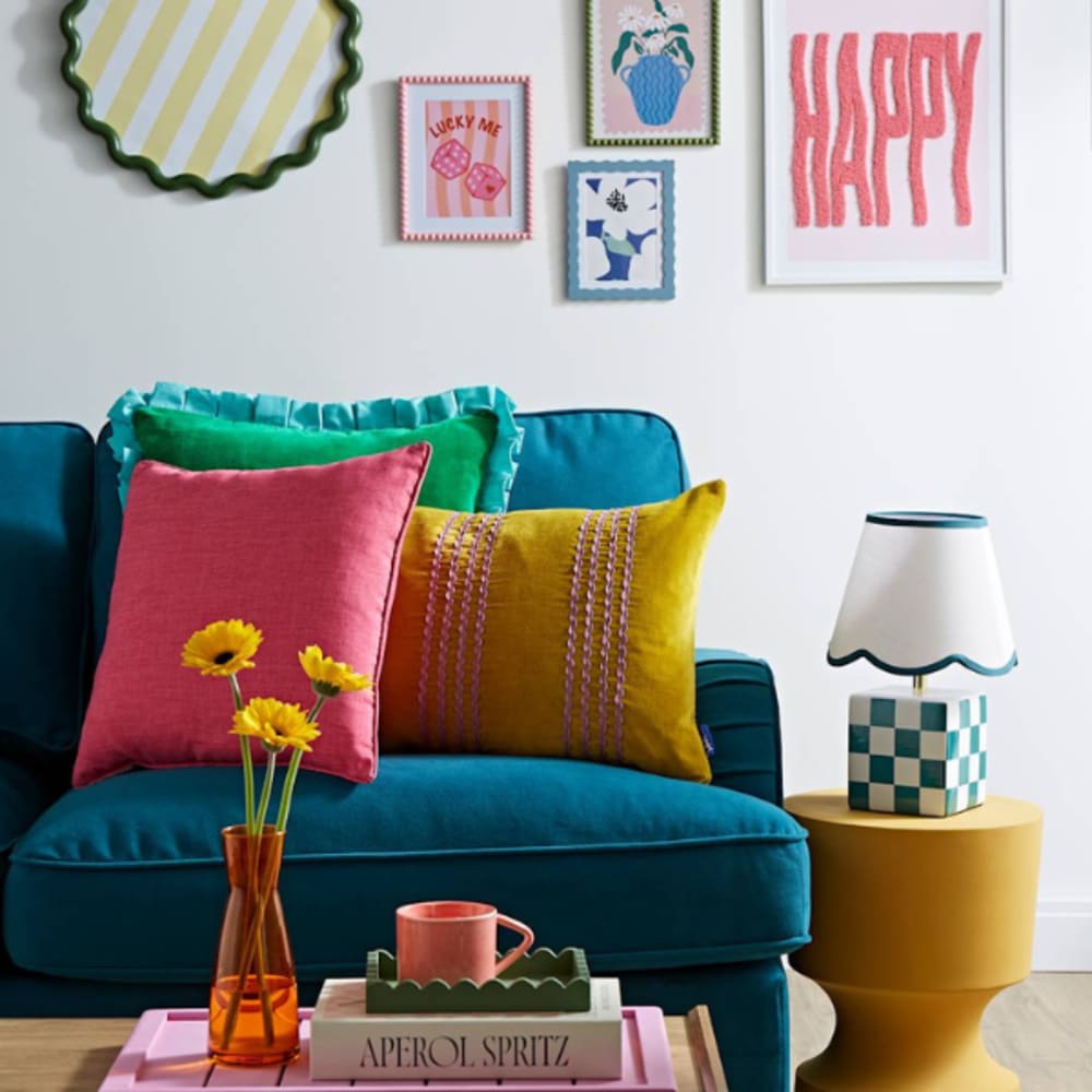 Modern colorful living room with teal sofa, bright cushions, yellow side table, checkered lamp, and eclectic wall art. Styled with vibrant décor accents and fresh flowers.