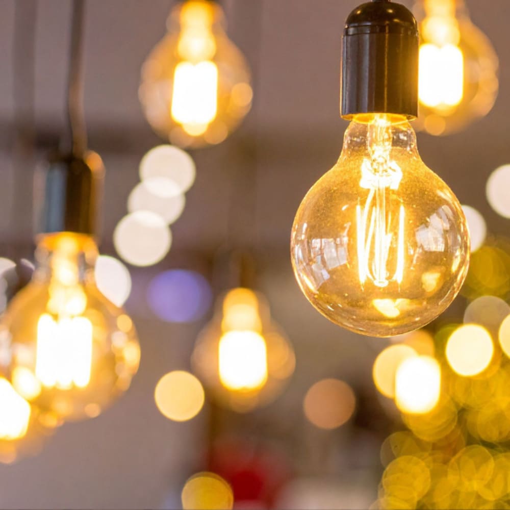 Close-up of warm glowing Edison-style filament light bulbs hanging from black cords, with a soft blurred background of additional lights creating a cosy, golden ambiance.