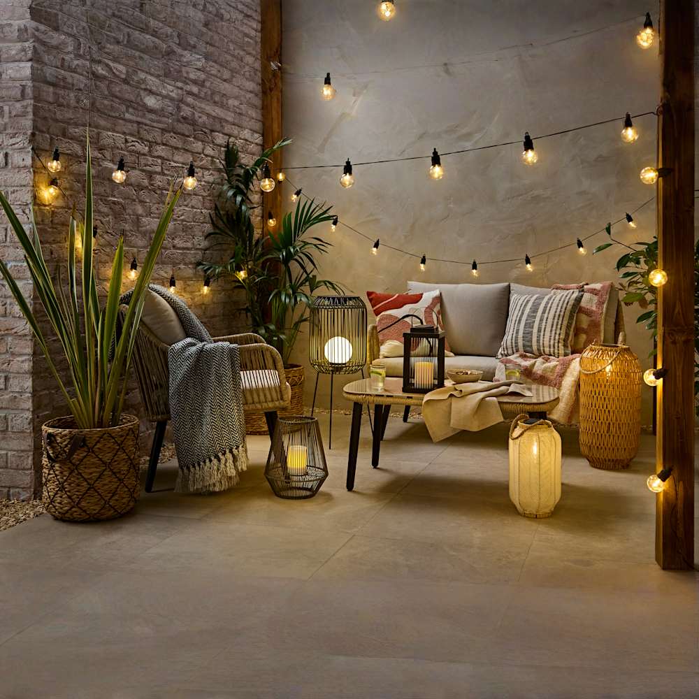 Outdoor patio styled with string lights, rattan furniture, and cosy cushions. Lanterns with LED candles and potted plants create a warm, inviting atmosphere for evening relaxation.
