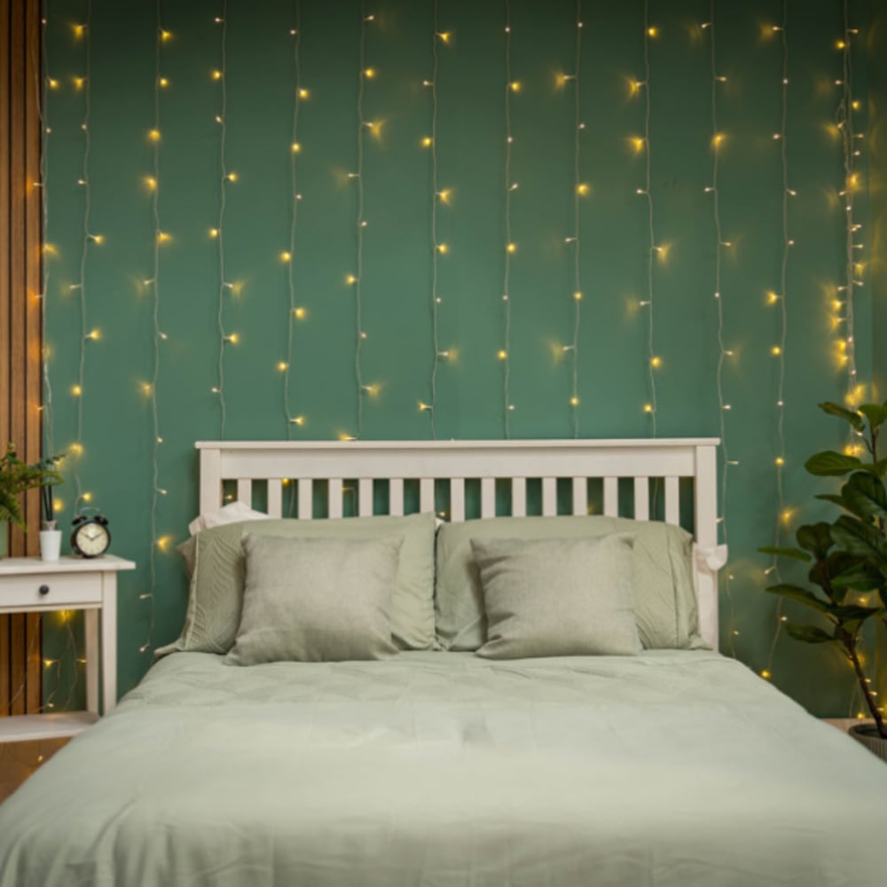 Cosy bedroom with a white wooden bed frame, soft green bedding, and a curtain of warm fairy lights hanging behind the headboard against a teal wall, creating a relaxing and inviting atmosphere.
