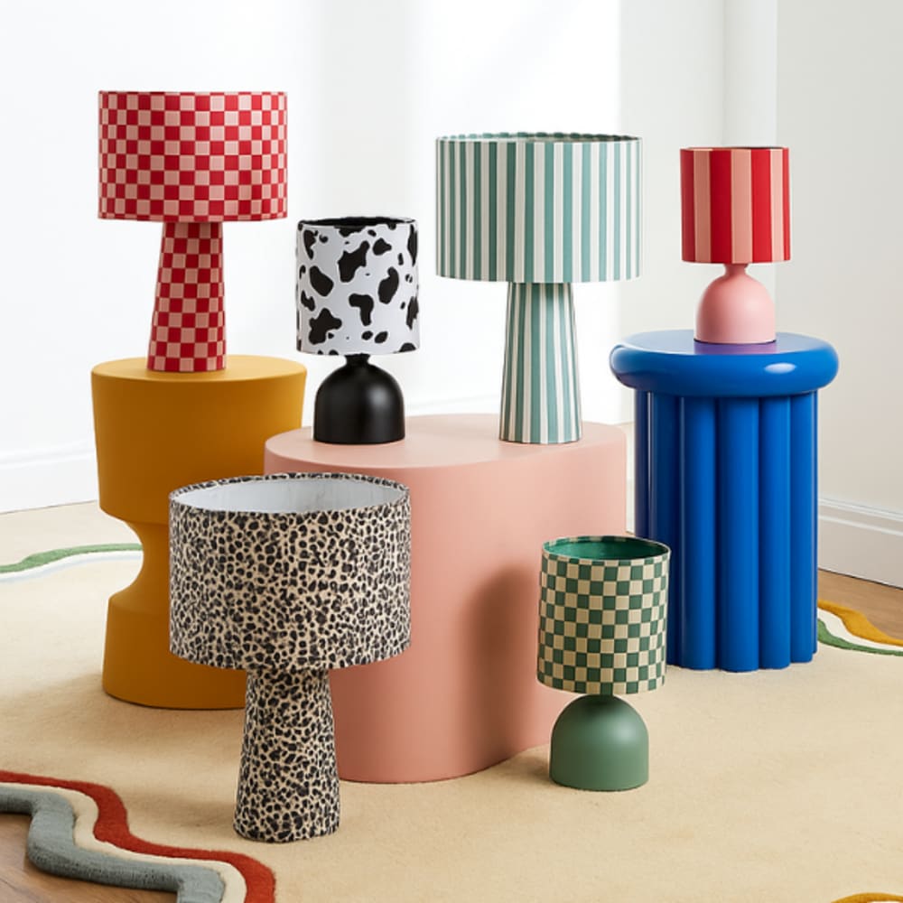 Collection of six bold and colorful table lamps with patterned shades, including red and green checkerboard, cow print, leopard print, and striped designs, displayed on sculptural side tables in a modern interior setting.
