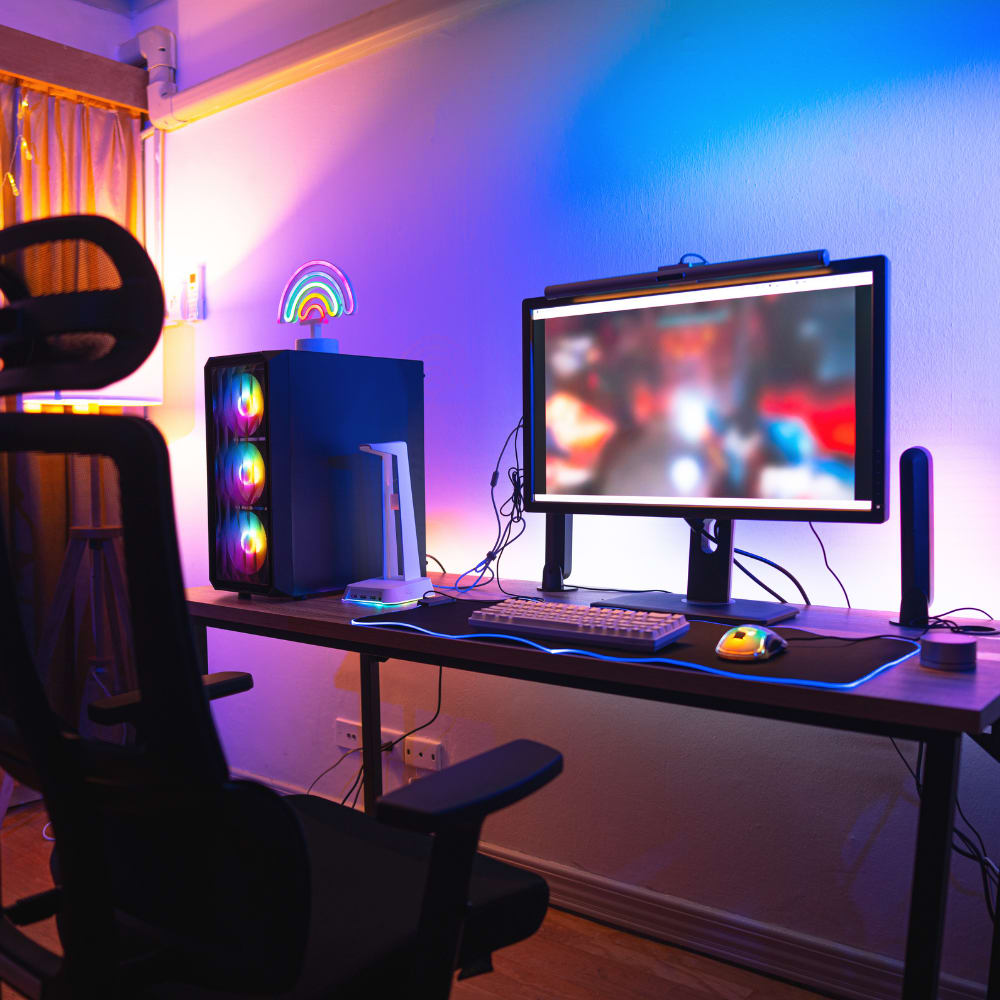 Stylish gaming desk setup with RGB PC tower, curved monitor, LED rainbow light, and ergonomic chair in a colourful ambient-lit room.
