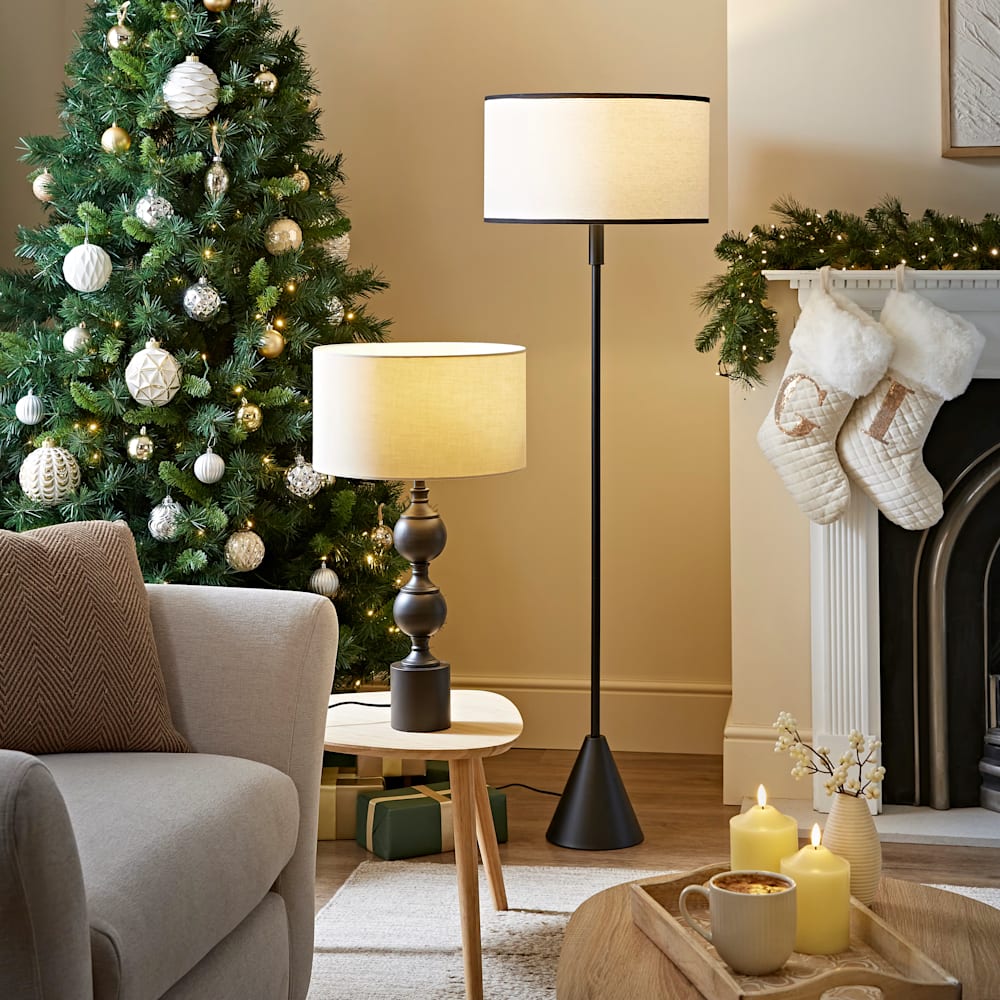 Cosy Christmas living room with a decorated tree, warm table lamp and black floor lamp, stockings on the fireplace, candles and wrapped gifts creating a snug festive atmosphere.