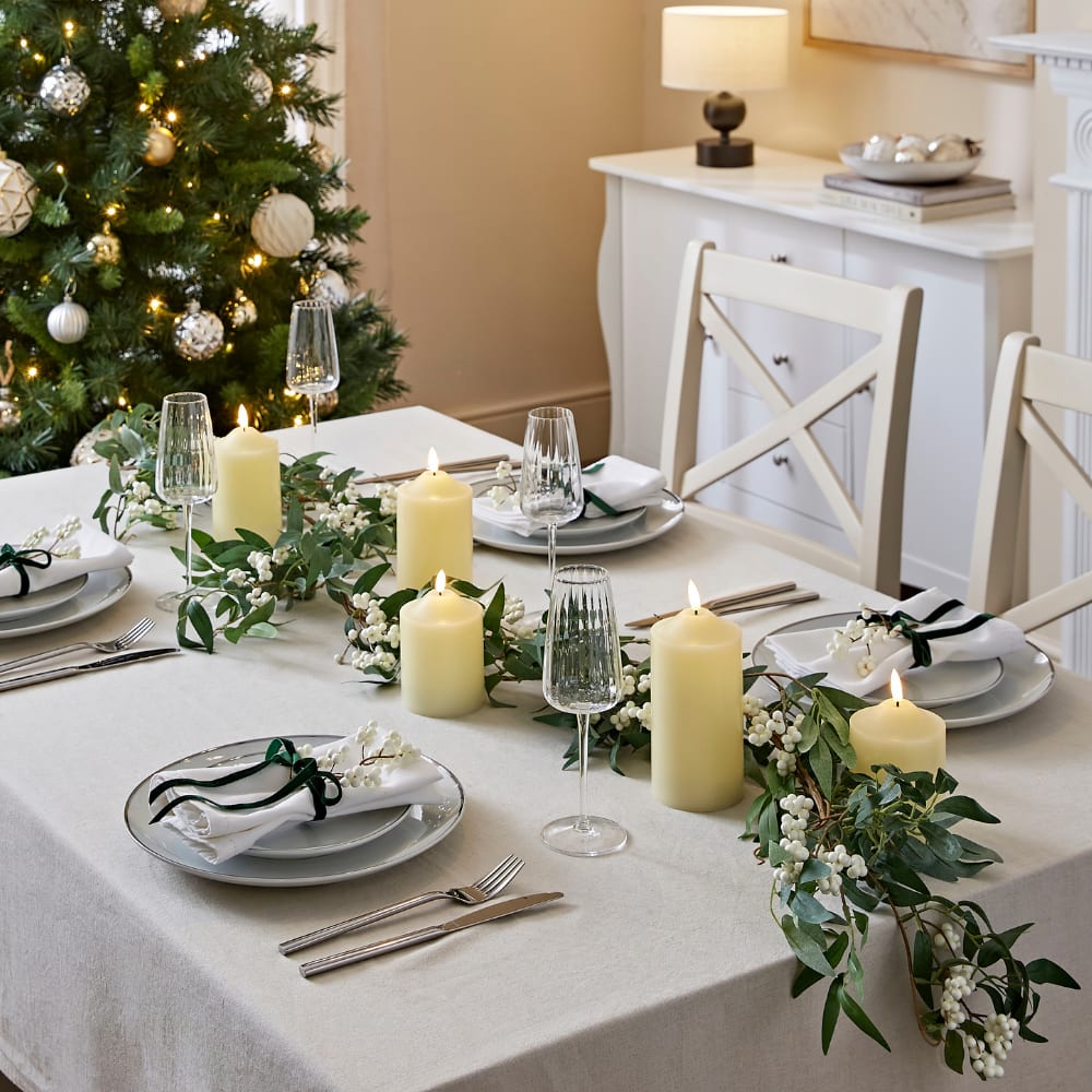Festive Christmas dining table with candles, greenery garland, elegant place settings, and glowing tree lights in a cosy neutral room