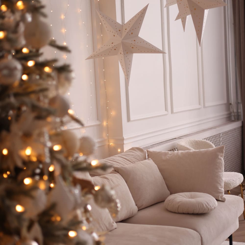 Cosy neutral living room with a beige sofa, glowing Christmas tree and decorative paper stars hanging above, styled with soft warm fairy lights for a festive ambience.