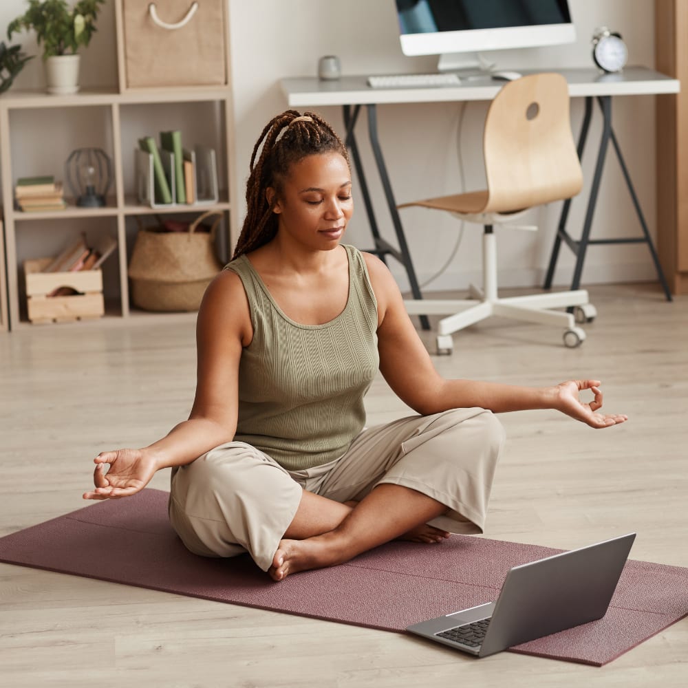 Woman meditating on a yoga mat at home while following an online yoga class on a laptop