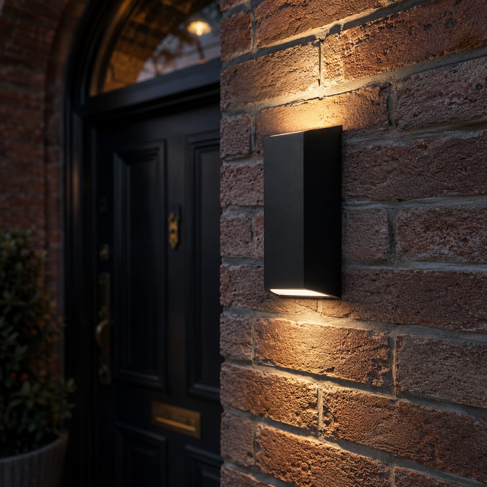 Up/down matte black wall light mounted on a brick wall beside a front door, casting soft light both upward and downward across the brickwork.