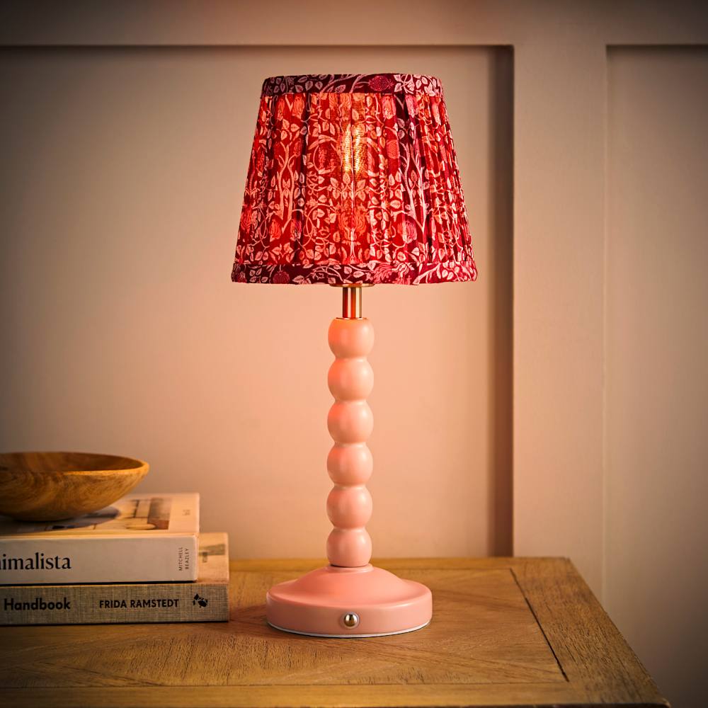 Pink rechargeable table lamp with a dark pink floral patterned fabric shade on a wooden side table, styled with books and warm ambient lighting.