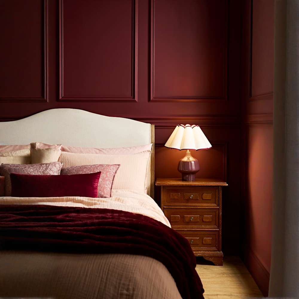 Elegant burgundy bedroom with cream upholstered bed, pink and velvet cushions, wooden bedside table and warm bedside lamp against dark wood panelled walls.